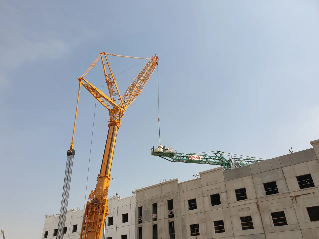 Yellow luffing-jib crane assembling a tower crane beside a concrete frame